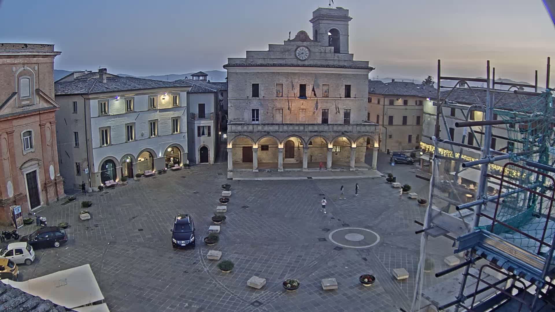 A town square at dusk, under a clear sky, features a large stone building with arched colonnades, several other buildings, a few cars, and some pedestrians.