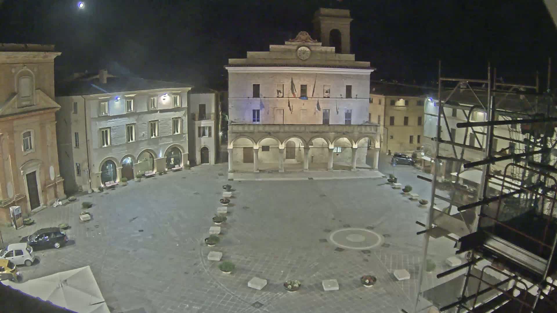 A nighttime view of a town square, featuring a large building with arches, smaller surrounding buildings, and a few cars parked near the edges, under a clear moonlit sky.