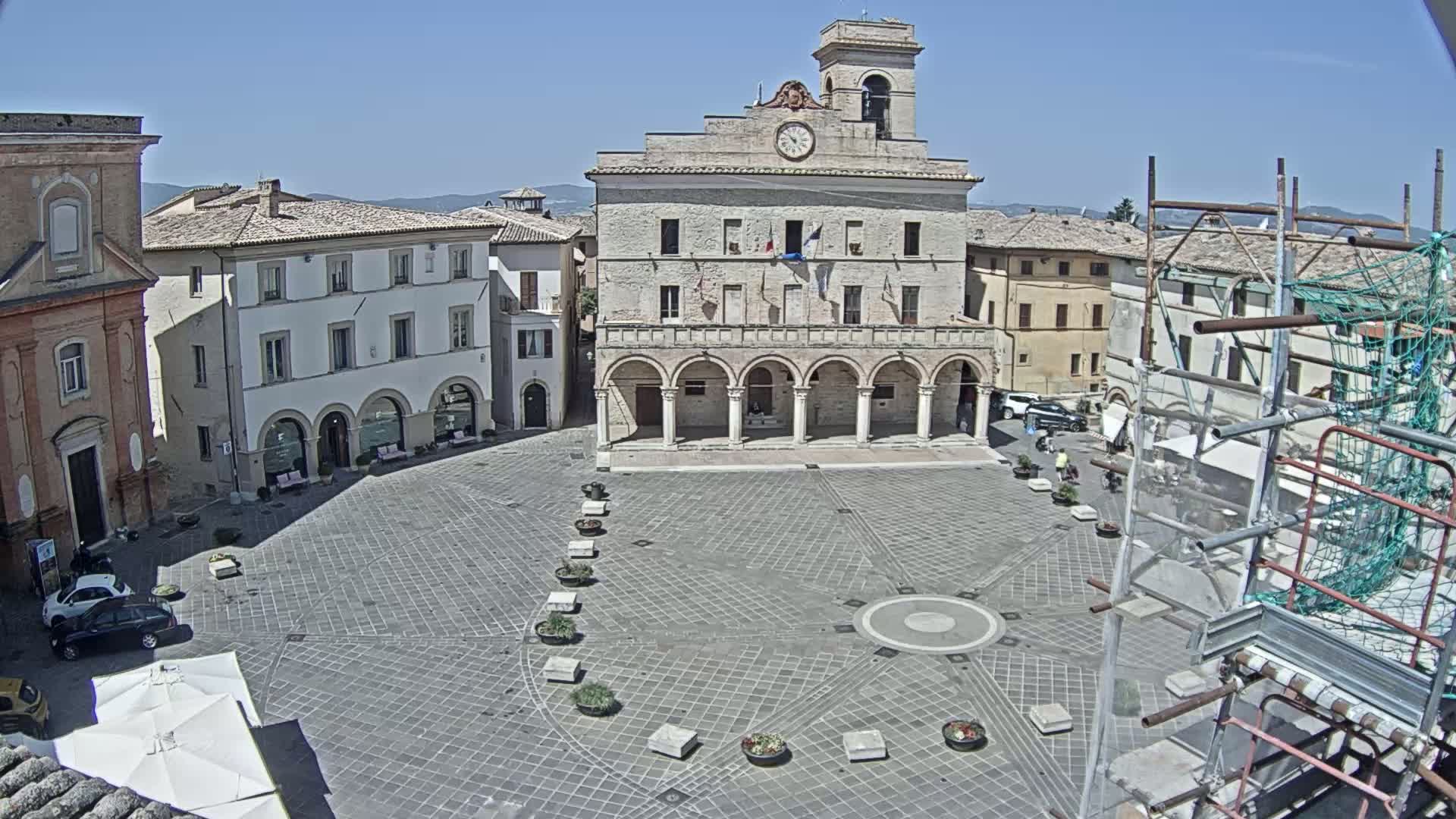 A large, stone plaza in a town features a prominent building with arched entryways, surrounded by other buildings, under a clear, sunny sky.