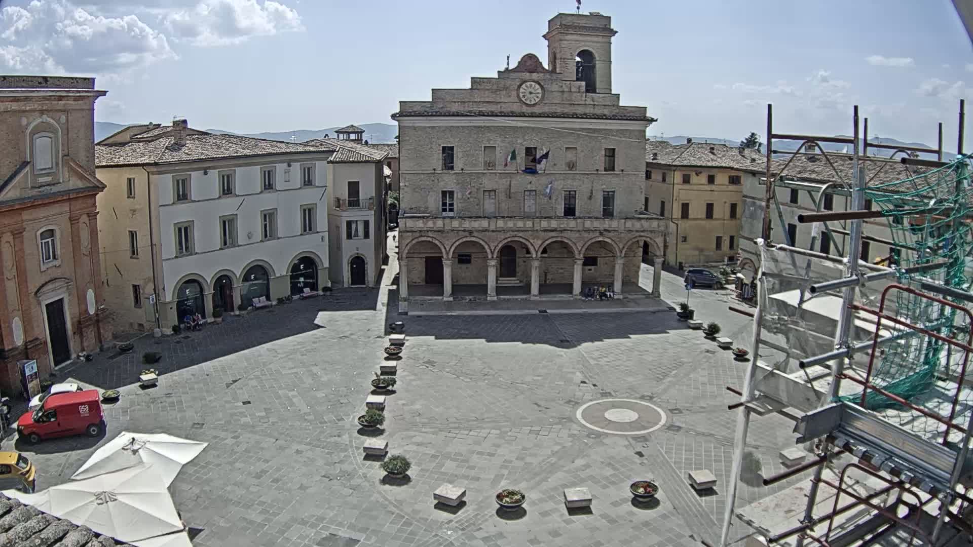 A town square with a large building featuring arched entryways and a clock tower is shown under sunny skies, with scaffolding visible on the right.