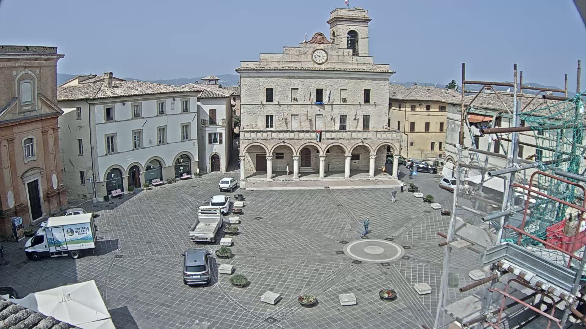 A large, stone town square with a clock tower in the center, surrounded by buildings, under a clear, sunny sky.