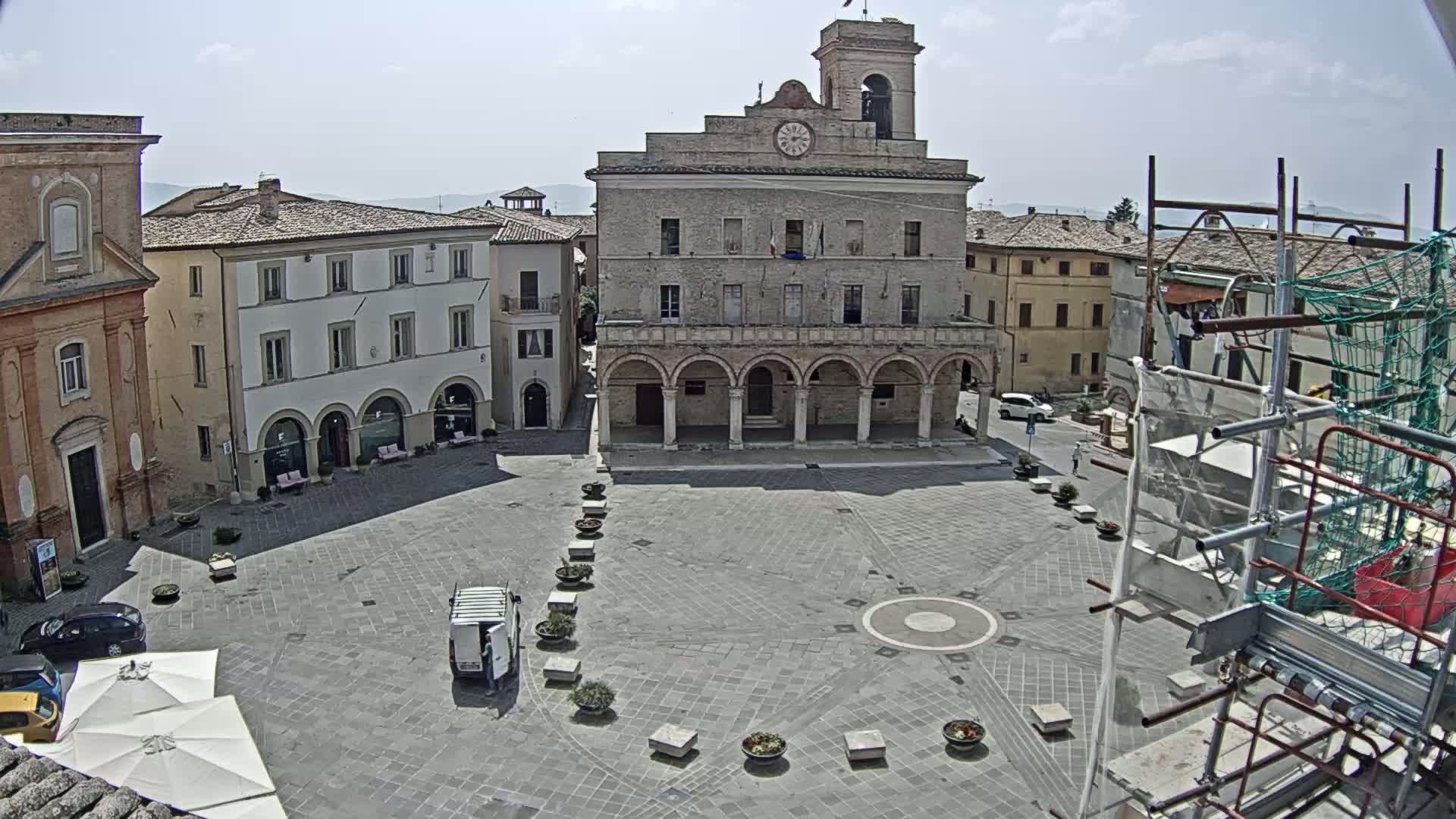 A large town square, under sunny skies, is centered around a large building with arched entryways, surrounded by other buildings and some scaffolding.