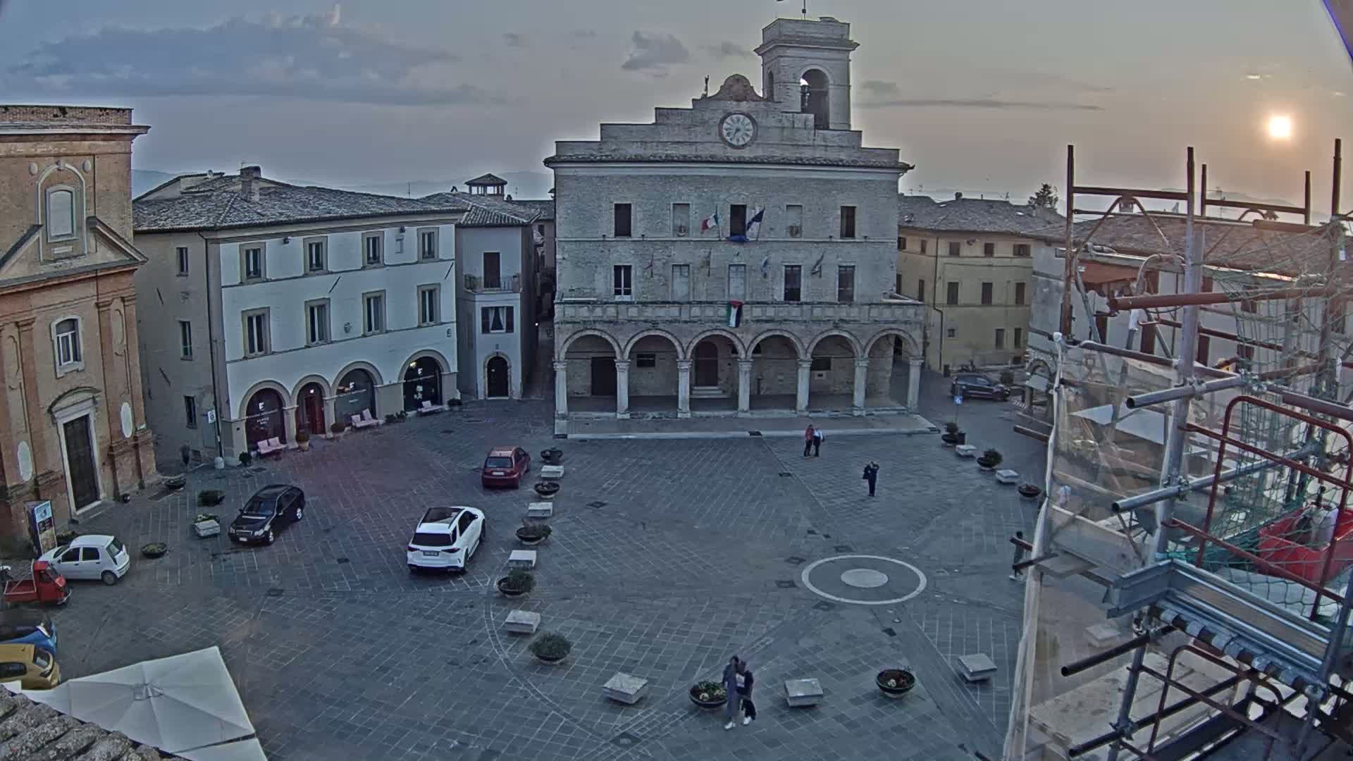 A town square with several cars parked, a large building with arches, and scaffolding, under a partly sunny sky.