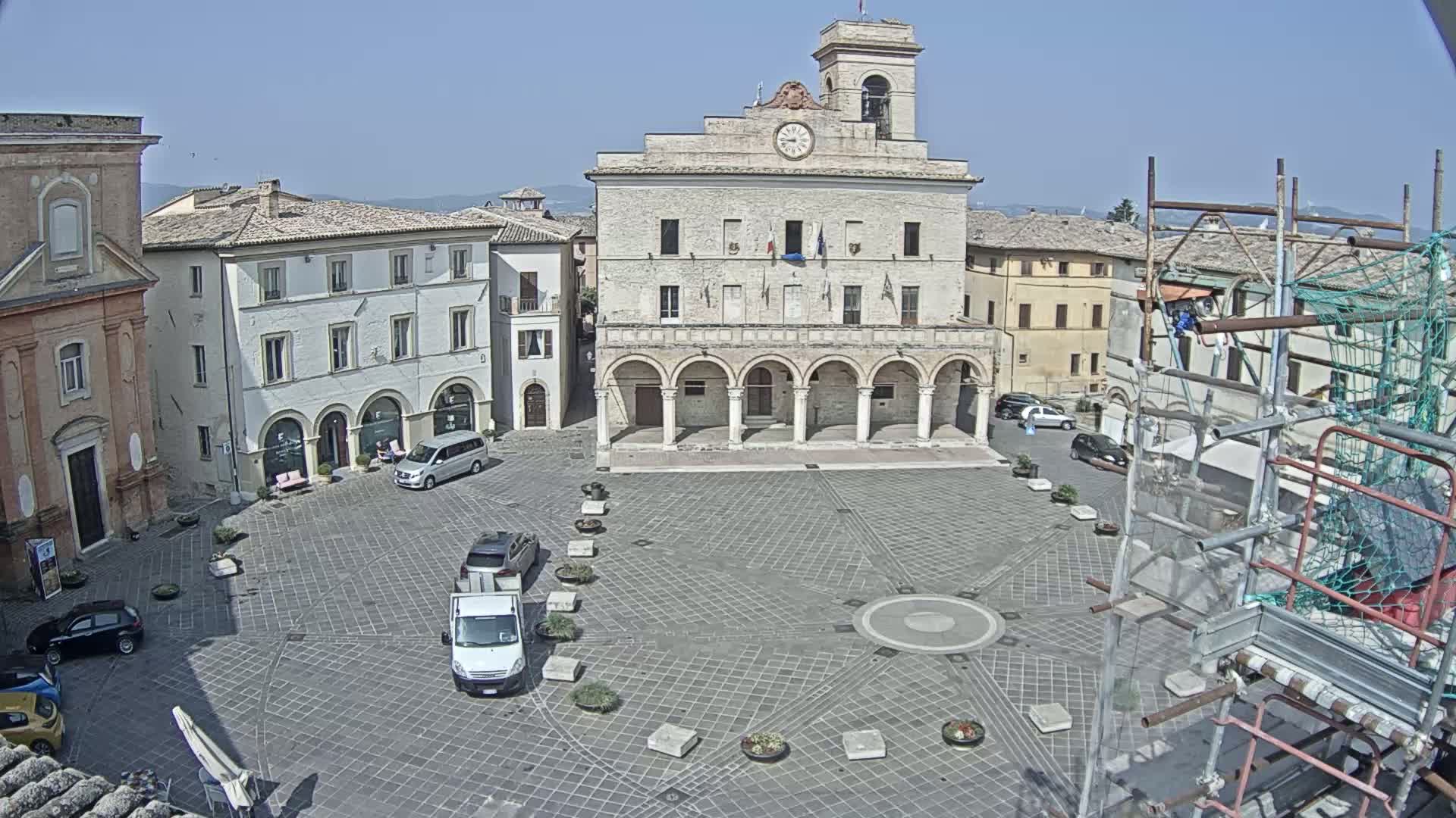 A large town square with a stone building, several parked cars, and scaffolding on the right, under a clear sky.