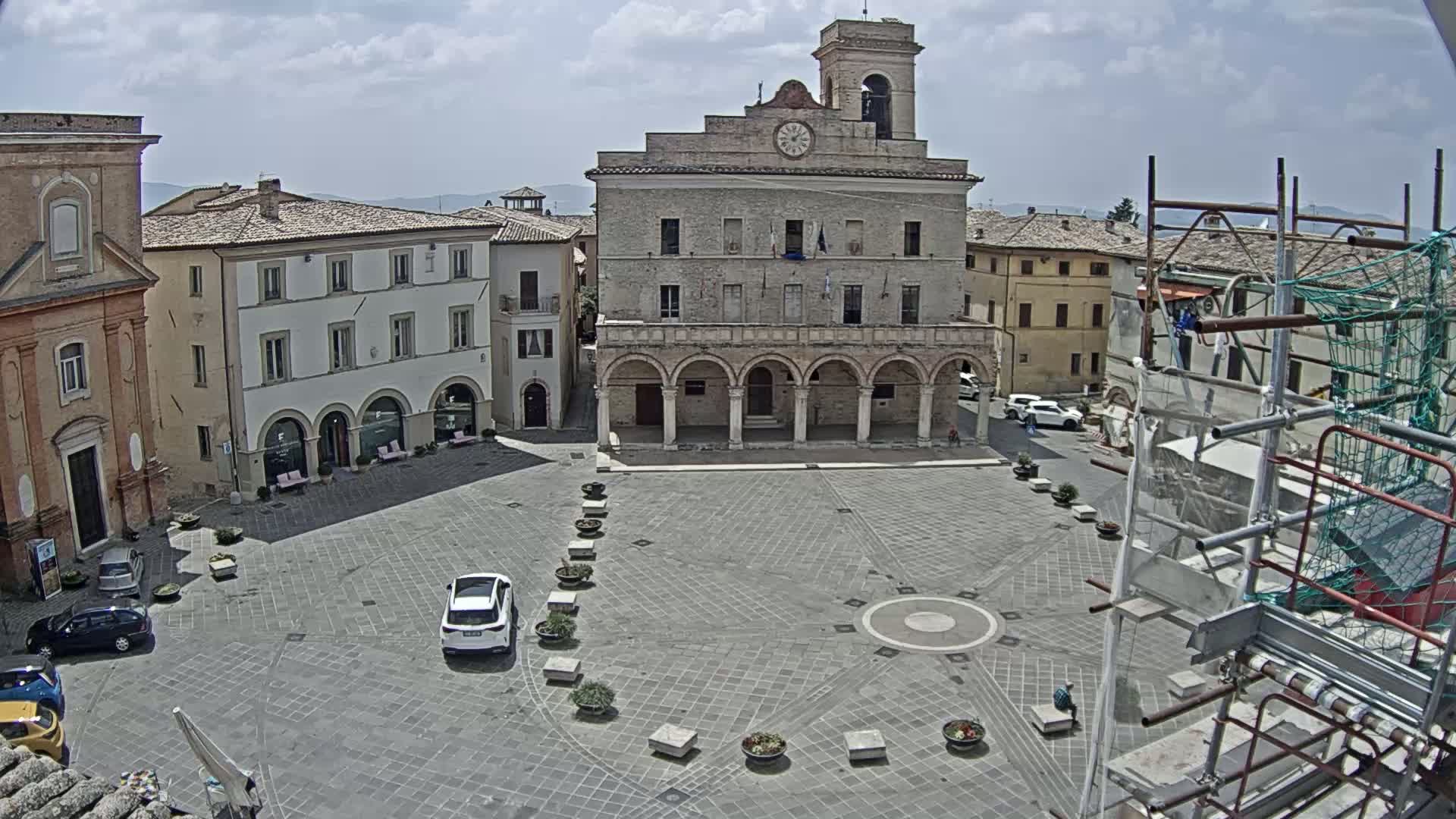 A town square with a large stone building featuring arched entryways, surrounded by other buildings, under a partly cloudy sky.