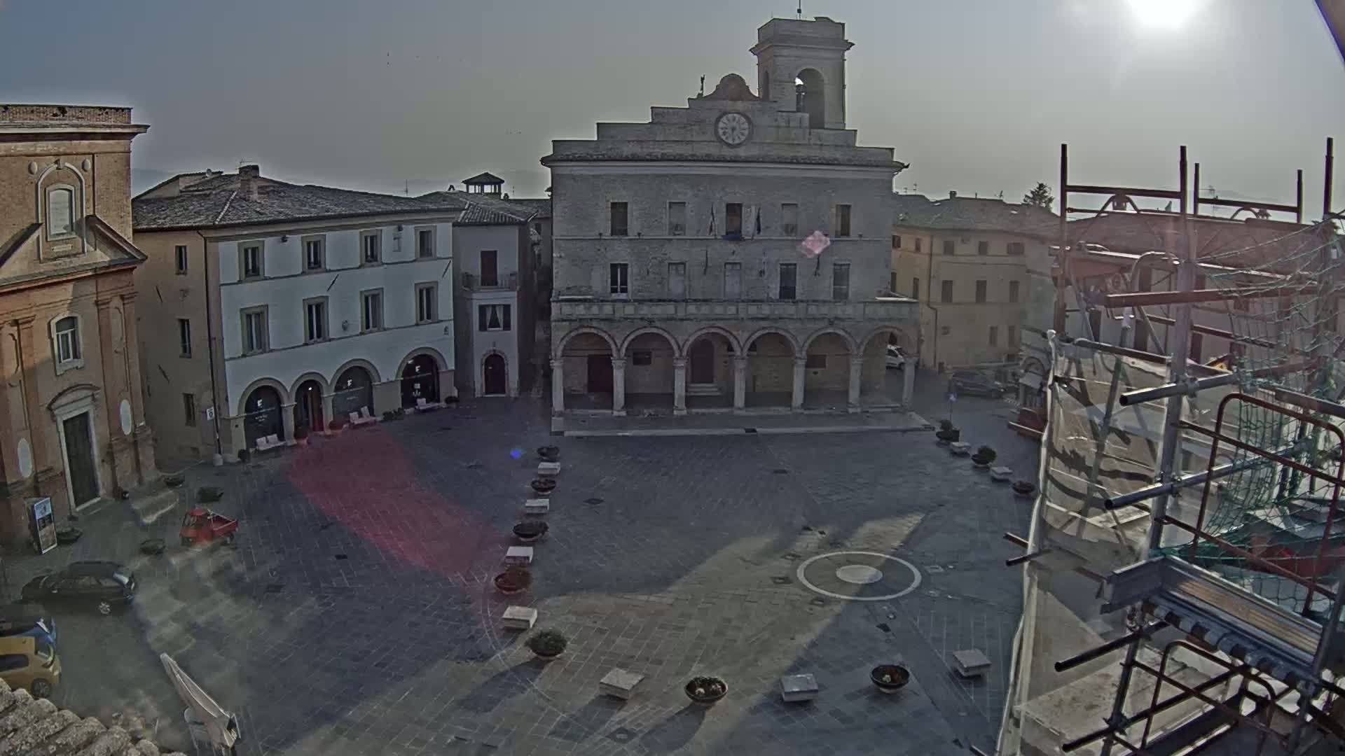 A mostly empty town square is viewed from above on a sunny, hazy day, centered on a large building with arched entryways and a clock tower, with scaffolding visible to the right.