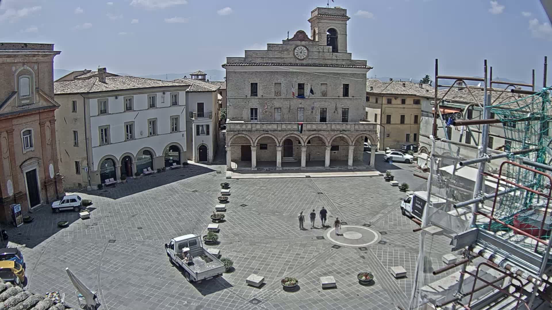 A town square with a large stone building, surrounded by other buildings, under a mostly sunny sky.