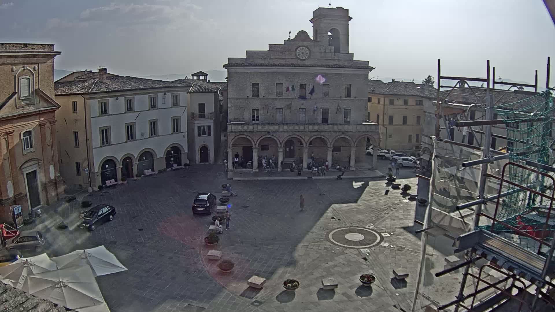 A town square with a large building and several cars, under a partly sunny sky.