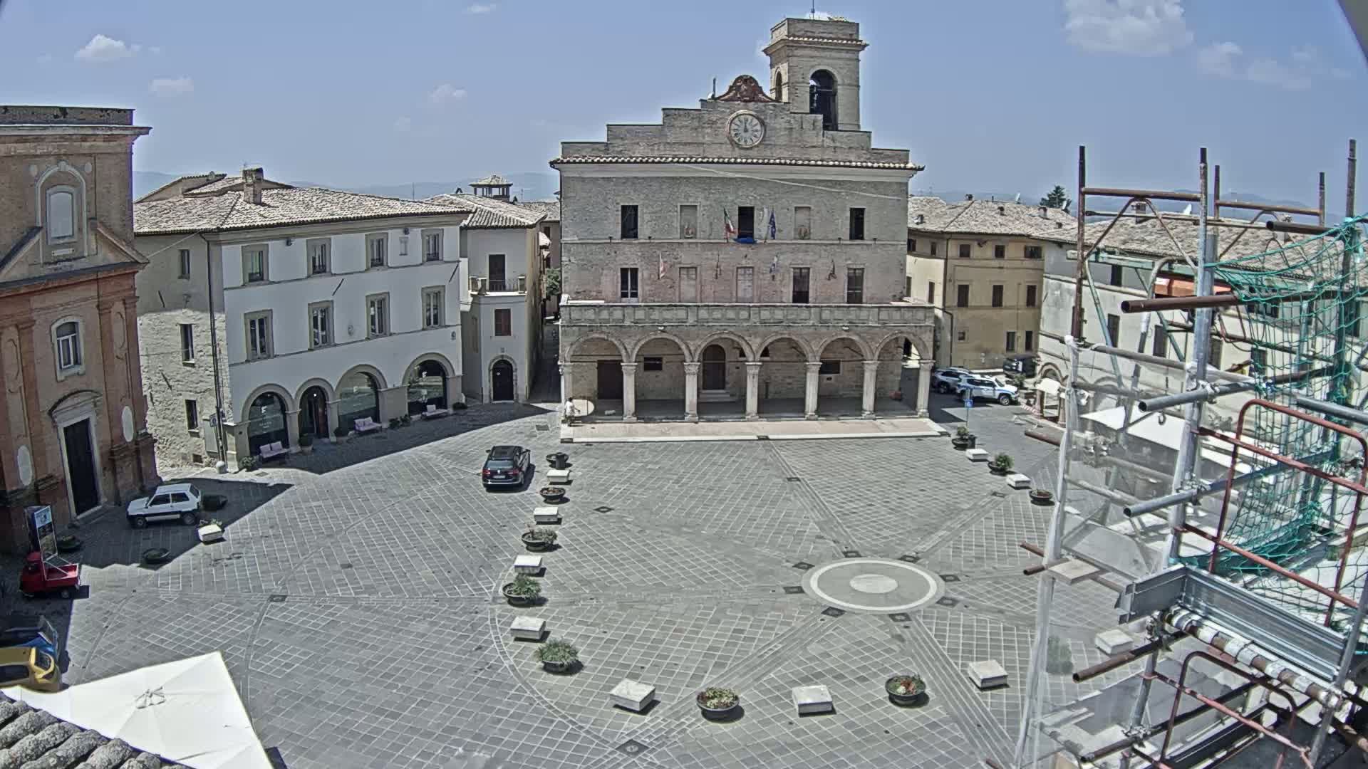 A large, paved town square is surrounded by historic buildings under a mostly clear, sunny sky, with scaffolding visible near one corner.