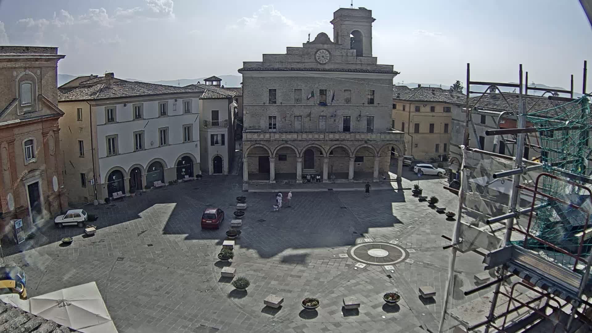 A sunny town square, centered on a large stone building with arched lower level, surrounded by other buildings, with a few cars and pedestrians visible.