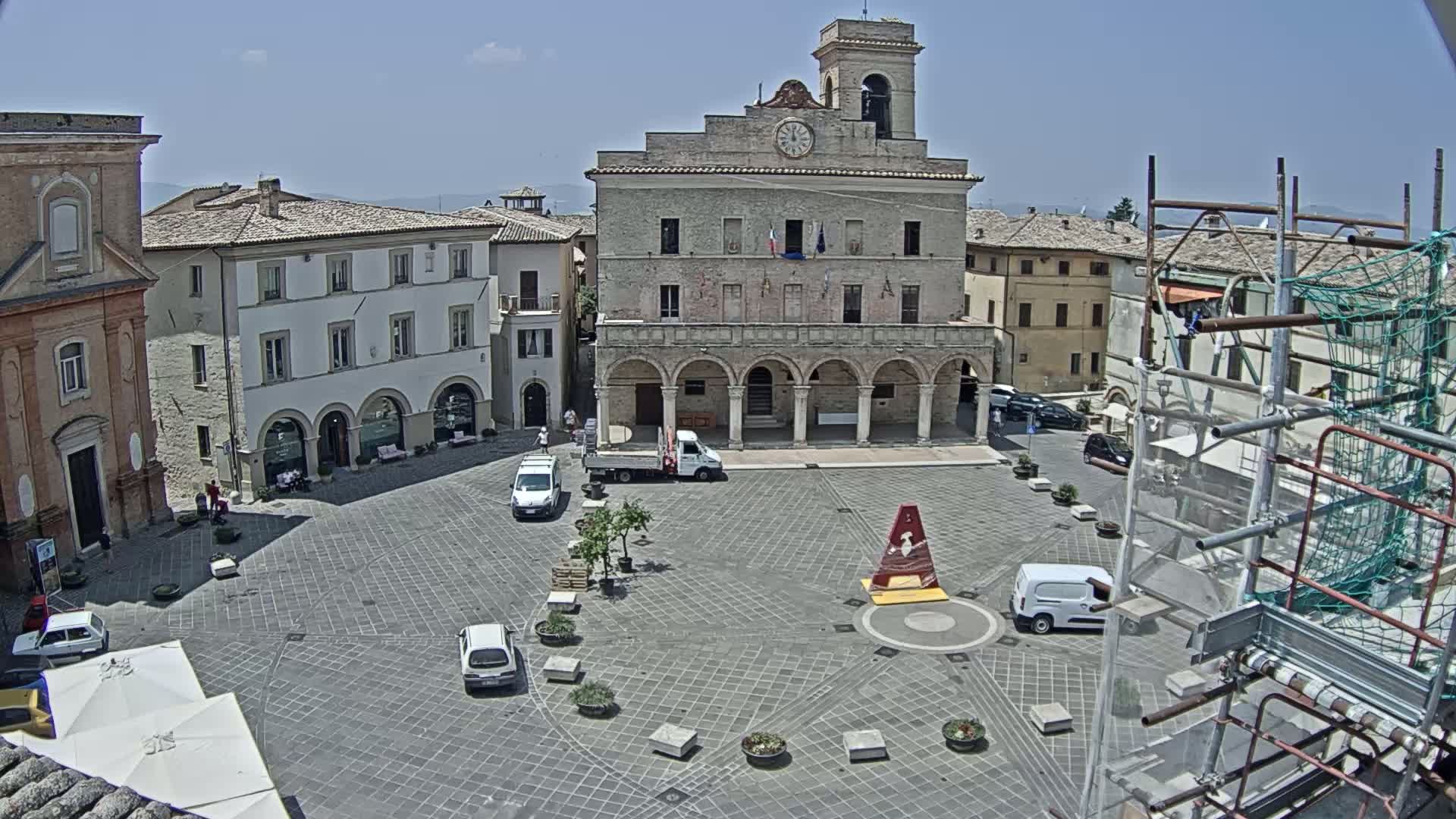A town square with a large stone building, several smaller buildings, and a few cars and pedestrians, under sunny skies.