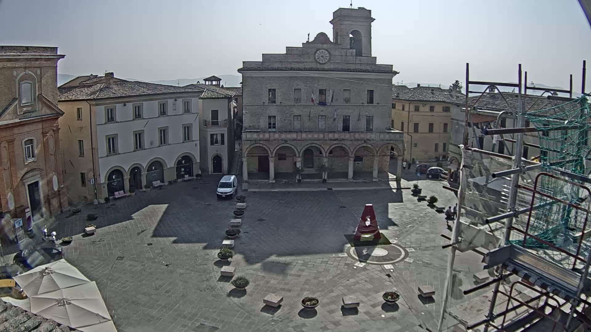 A town square with a large building featuring arched entryways is shown on a sunny day; scaffolding is visible to the right.
