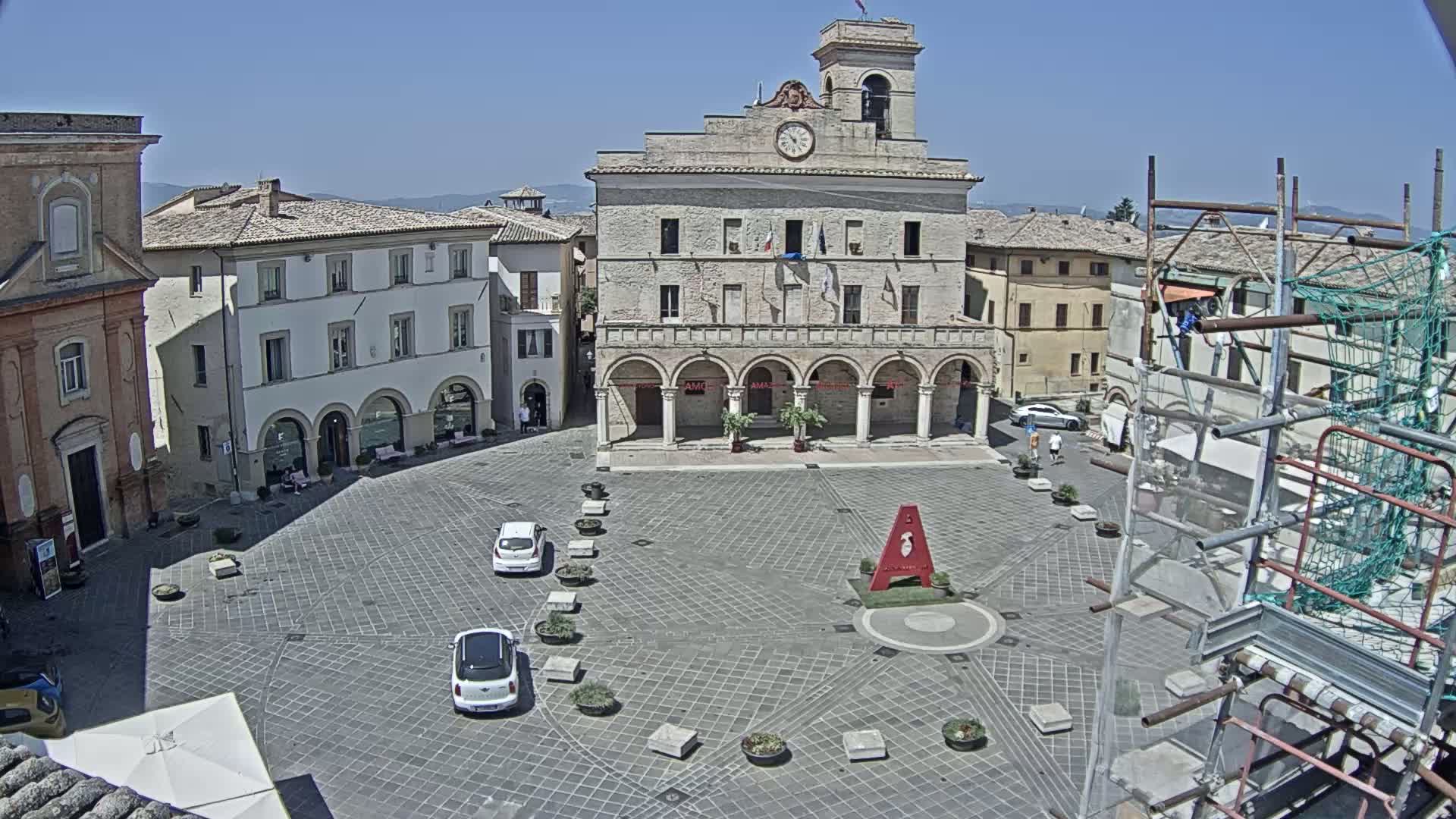 A large town square with a prominent stone building, several parked cars, and scaffolding is seen on a sunny day.