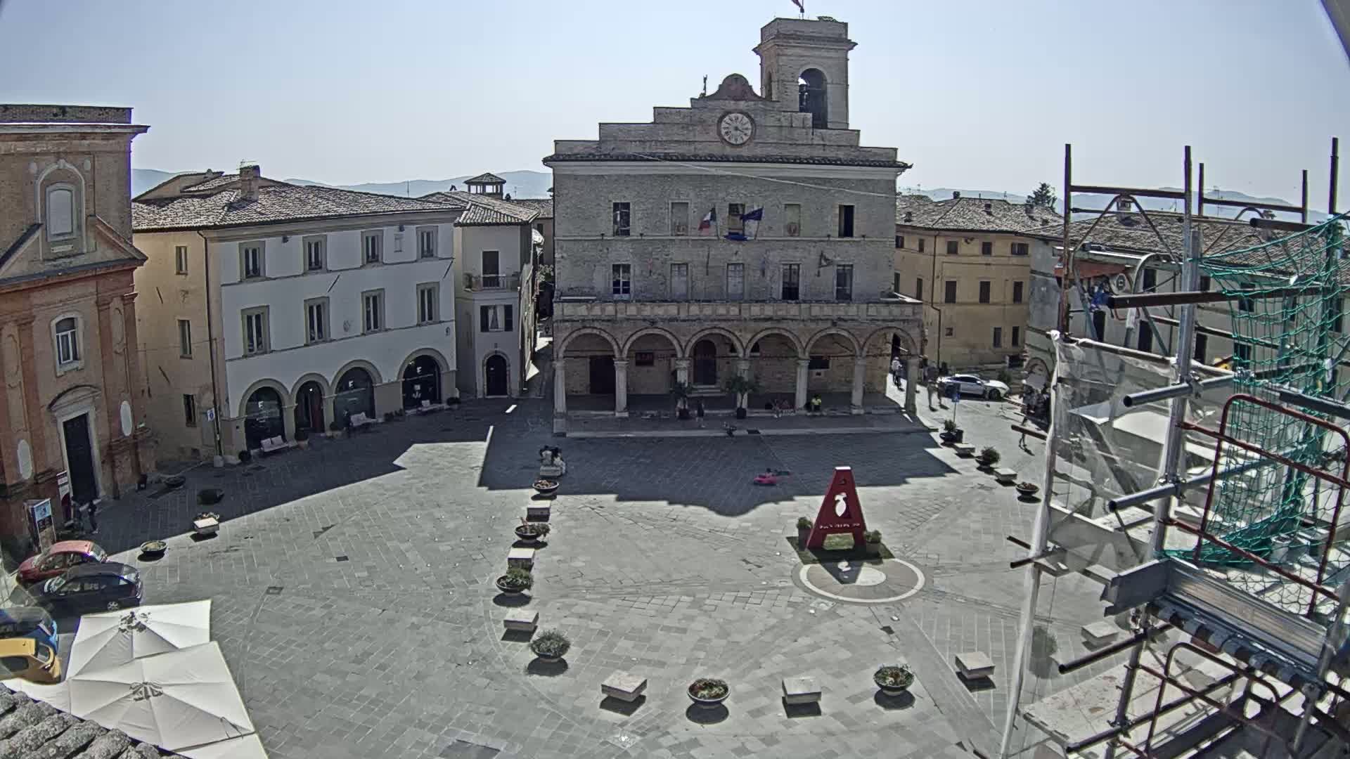 A large town square with a clock tower and surrounding buildings, under sunny skies.