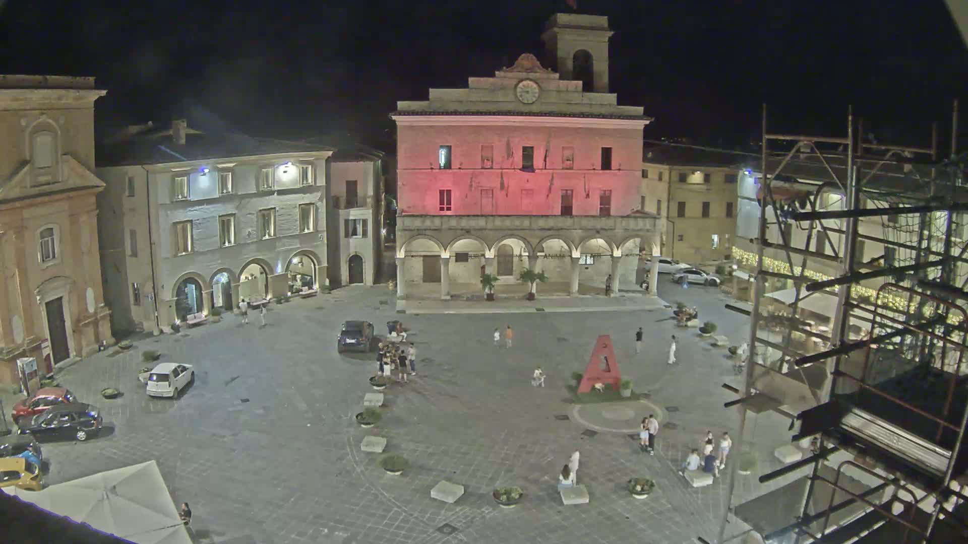 A large town square with a clock tower and surrounding buildings, under sunny skies.