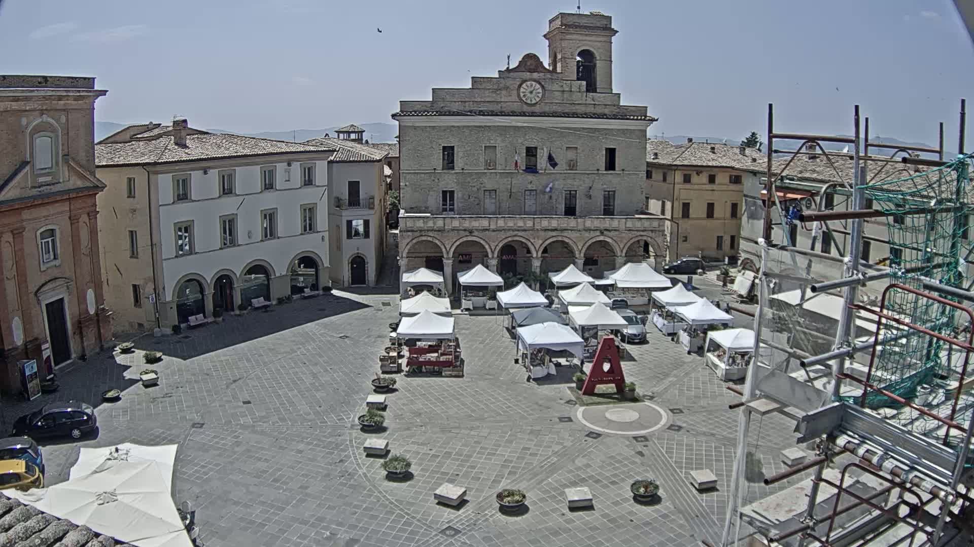 A town square with several white market stalls set up in front of a large stone building on a sunny day.