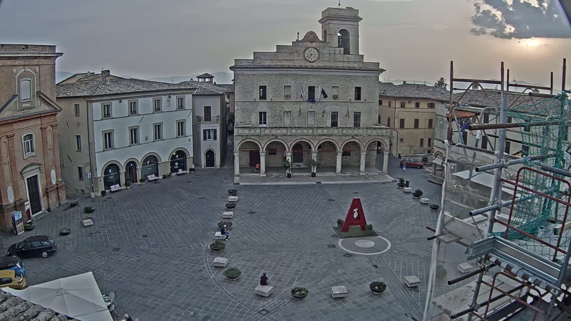 A large town square, under an overcast sky, is surrounded by historic buildings, one of which has a clock tower, with scaffolding visible in the lower right.
