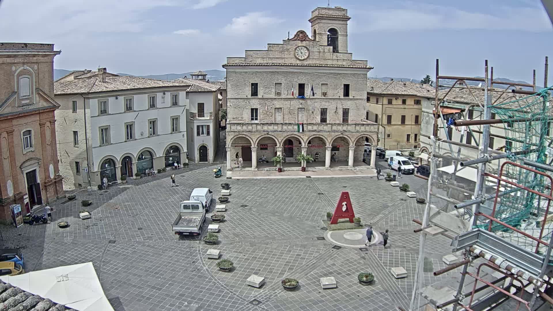 A town square with a large stone building, several smaller buildings, and a few vehicles under a partly cloudy sky.