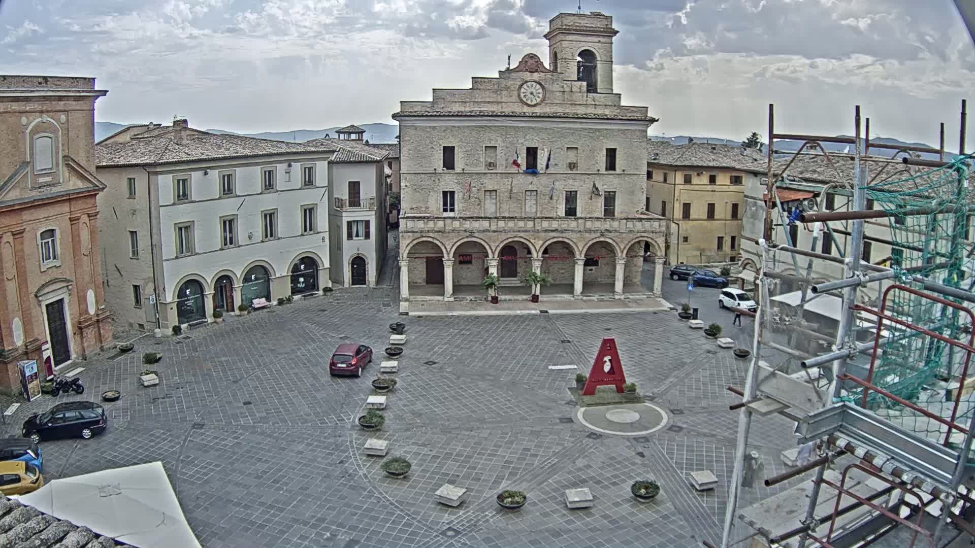 A town square with a large building, several smaller buildings, a few cars, and scaffolding is visible under partly cloudy skies.