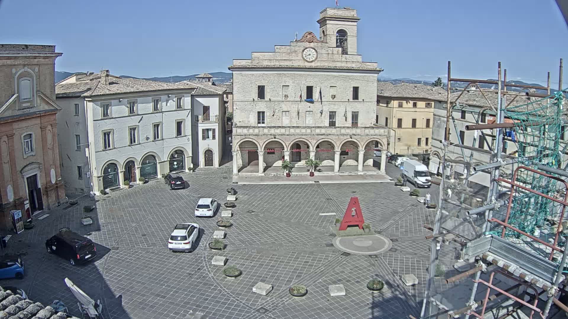 A town square with several cars parked, surrounded by buildings including a large town hall with arches, under a clear, sunny sky.