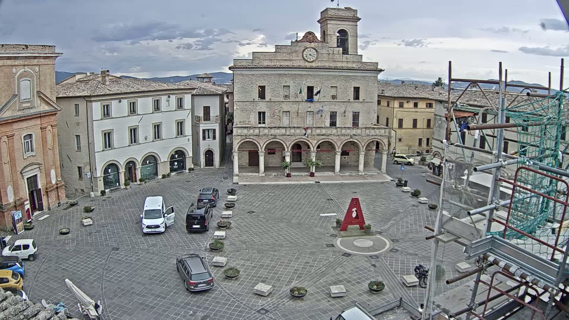 A town square with a large stone building, several parked cars, and scaffolding in the corner under a cloudy sky.