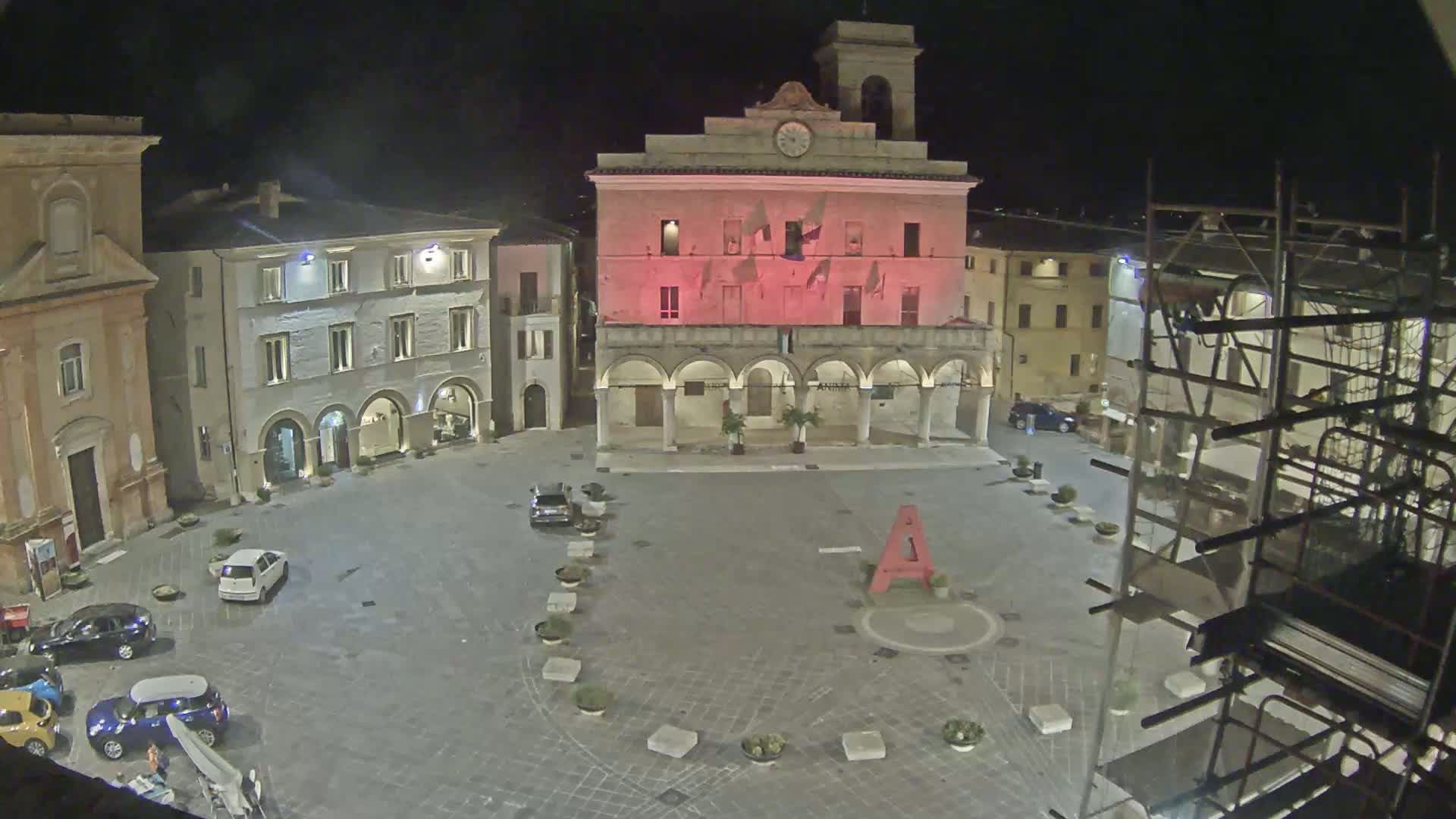 A nighttime view of a town square with several buildings, including a large building with arched entryways that is illuminated in pink, and several cars parked in the square.