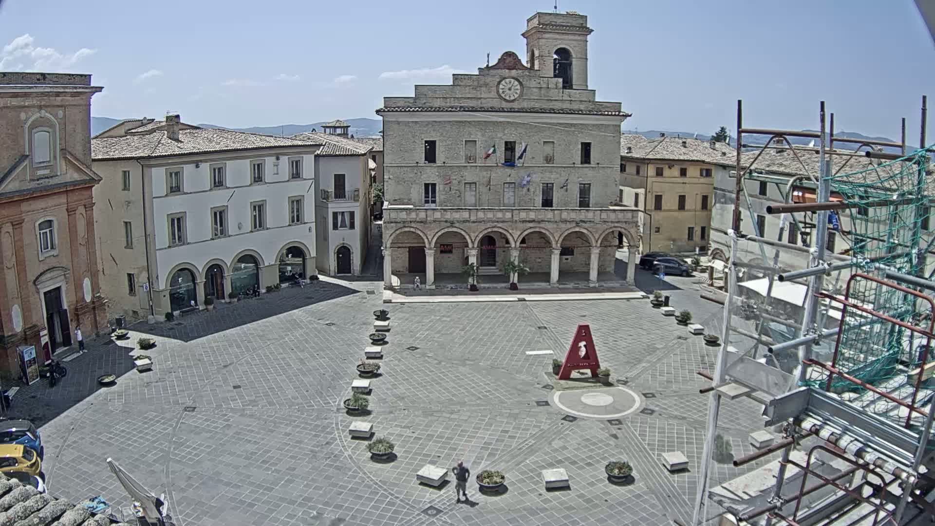 A town square with a large stone building in the center, surrounded by other buildings, under a sunny sky.