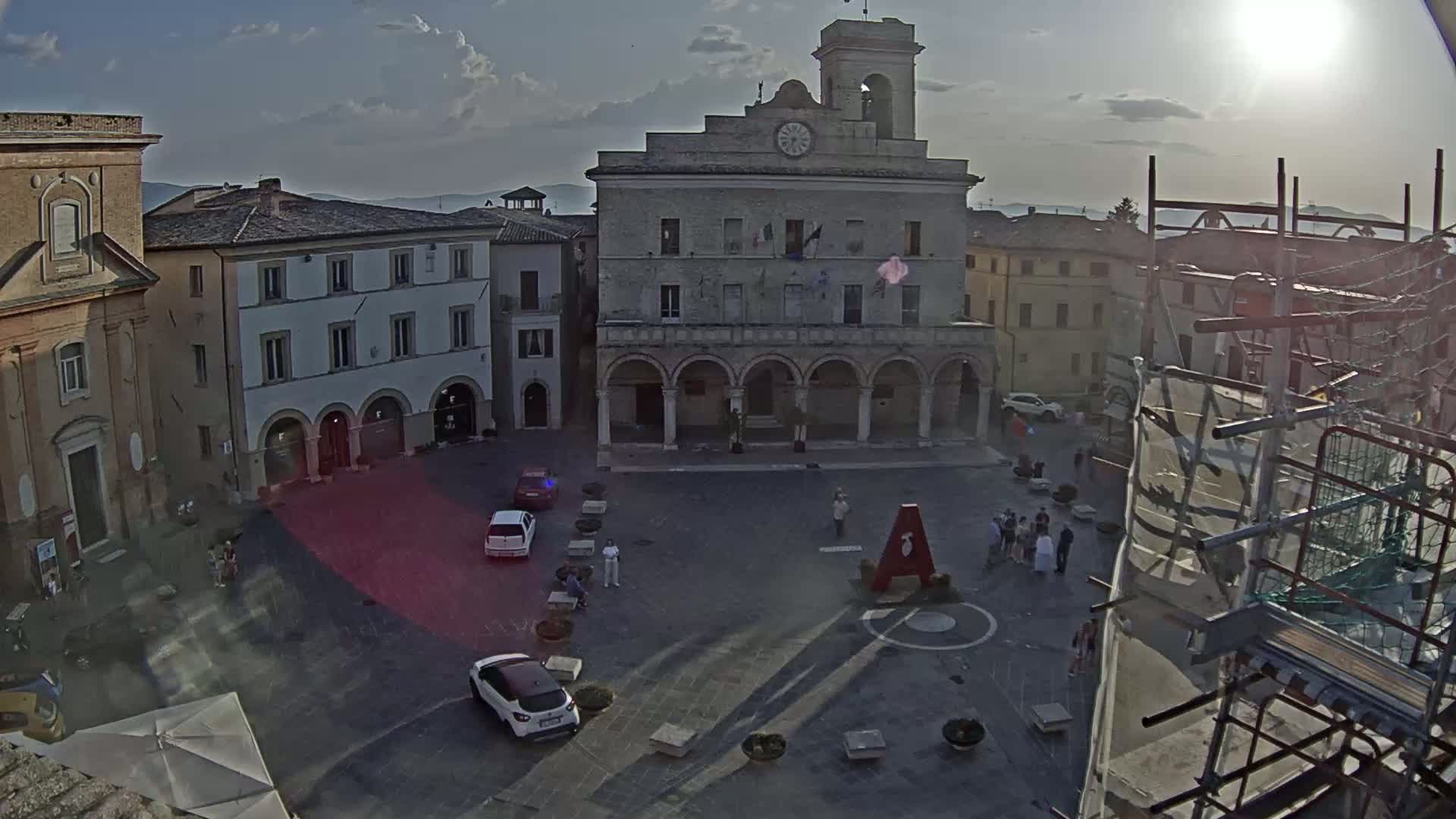 A sunny day reveals a town square with several buildings, a few cars, and people scattered throughout the area, construction scaffolding is visible to the right.