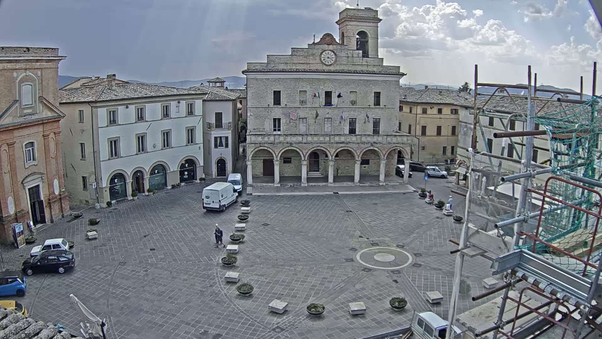 A large town square with a prominent stone building, several smaller buildings, a few cars, and some pedestrians, under partly cloudy skies.