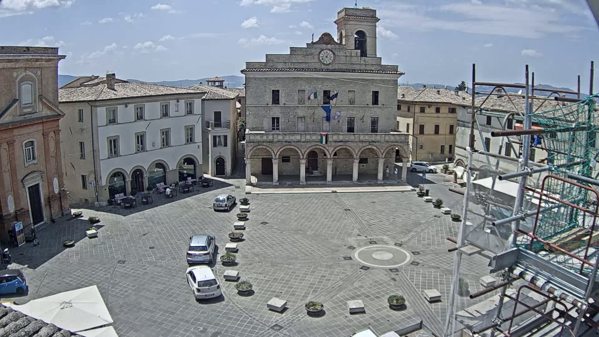 A town square with a large stone building, surrounded by other buildings, under a mostly sunny sky.