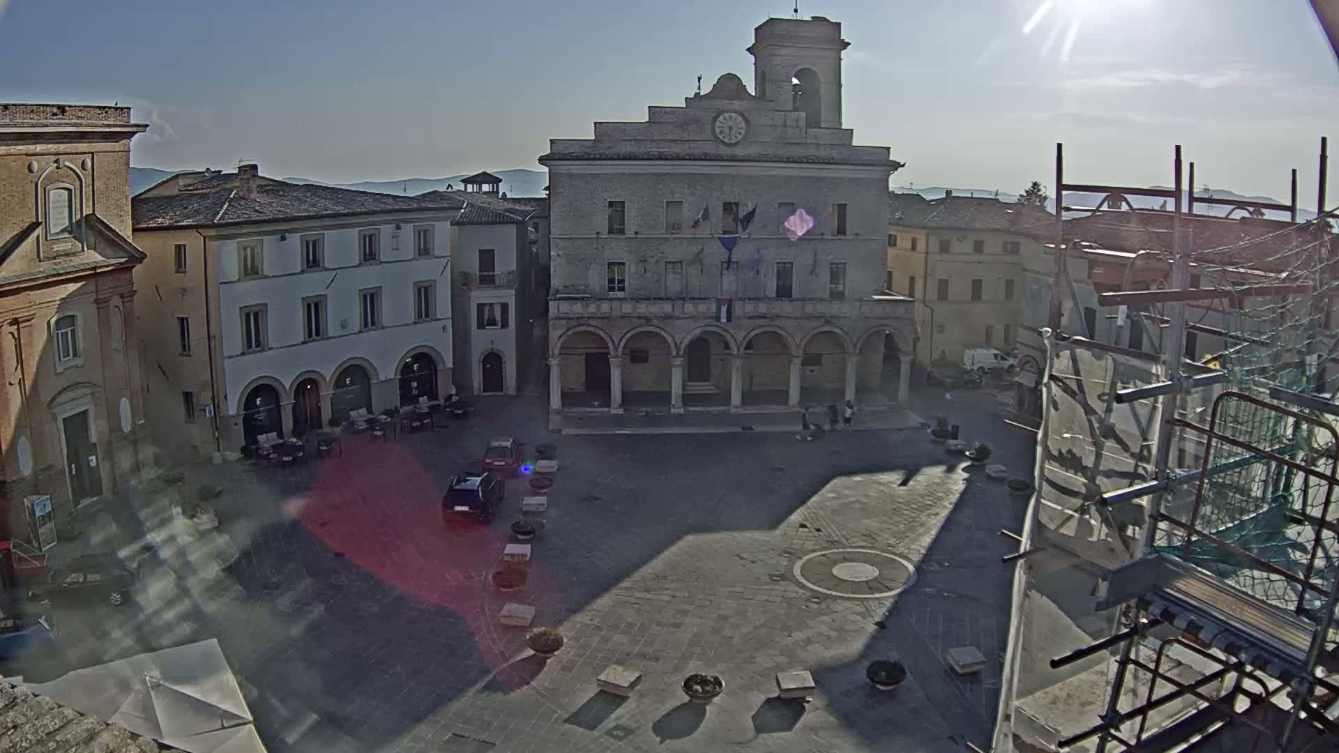 A sunlit town square, centered around a large, arcaded building, with several smaller buildings surrounding it under a clear sky.