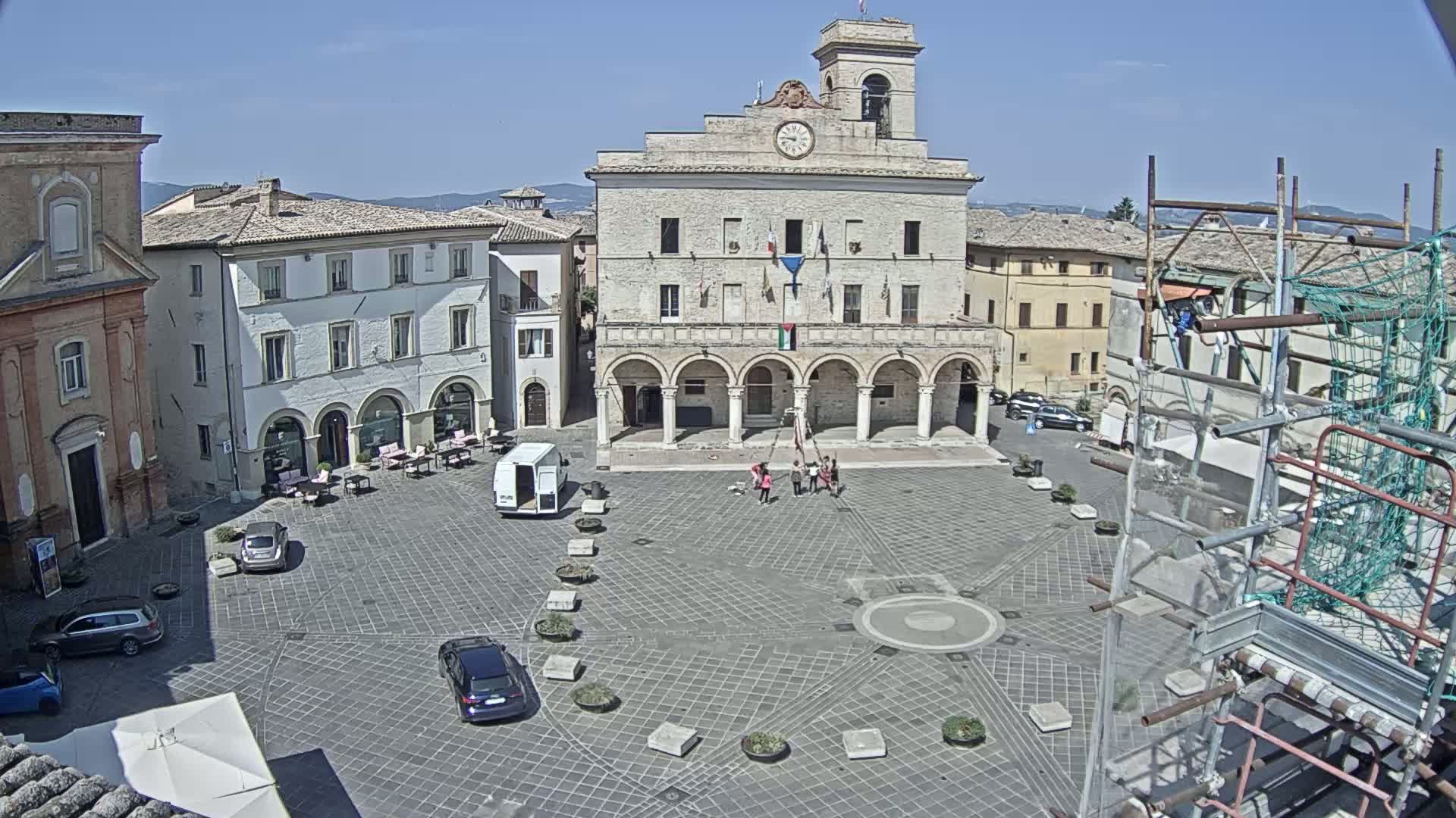 A town square with a large stone building in the center, several cars parked, a few people gathered, and scaffolding to one side, under sunny skies.
