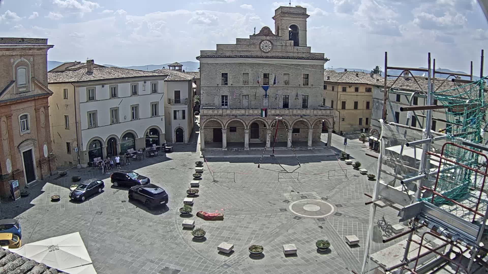 A town square with several buildings, a few cars, and scaffolding under a mostly sunny sky.