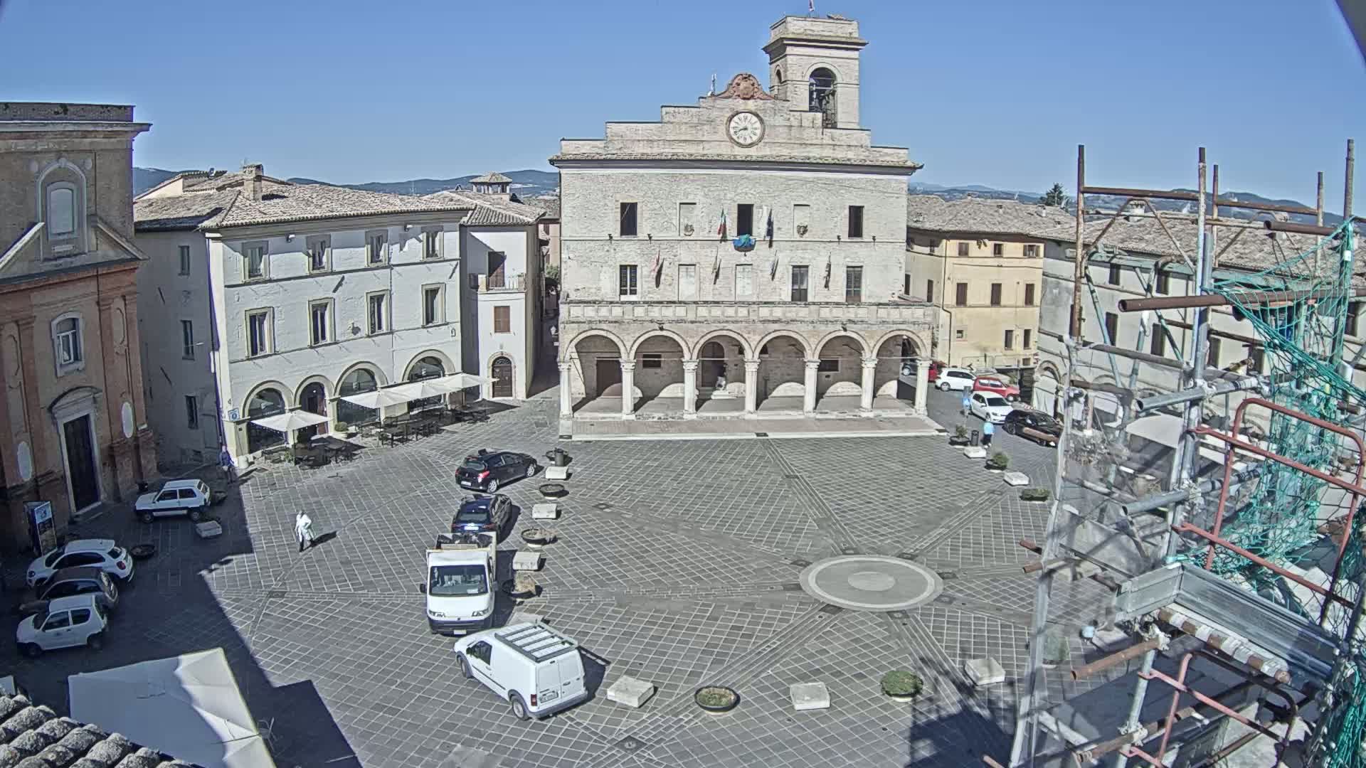 A town square with a large stone building, several parked cars, and scaffolding is visible on a sunny day.