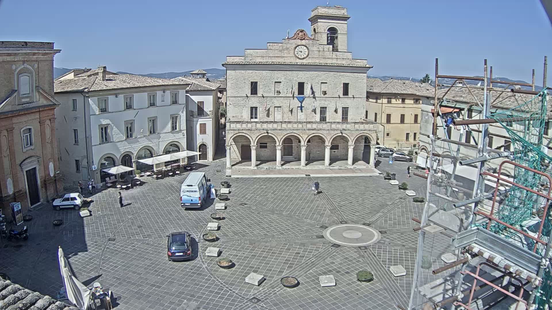 A sunny day reveals a town square centered on a large, arched building, surrounded by other buildings and scaffolding, with several cars and a few people visible.
