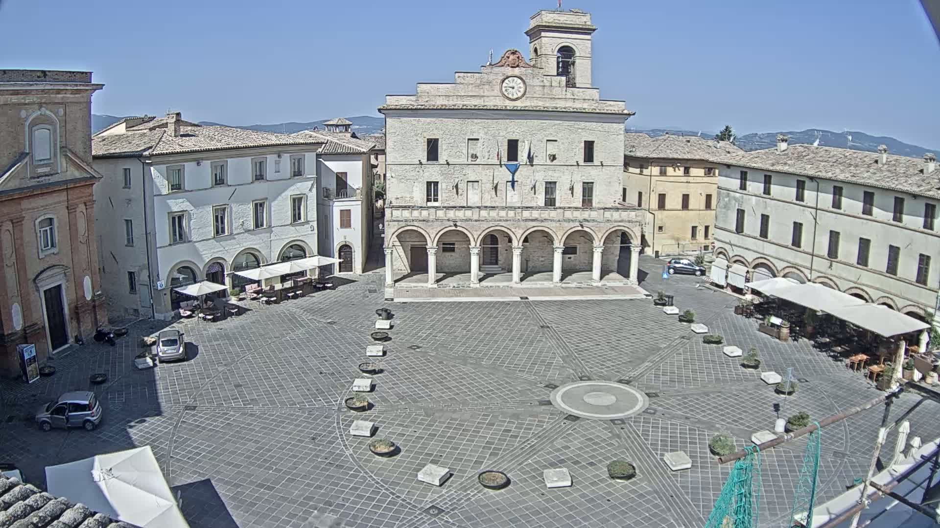 A large, paved town square is centered on a grand building with arched entryways, under a clear blue sky.