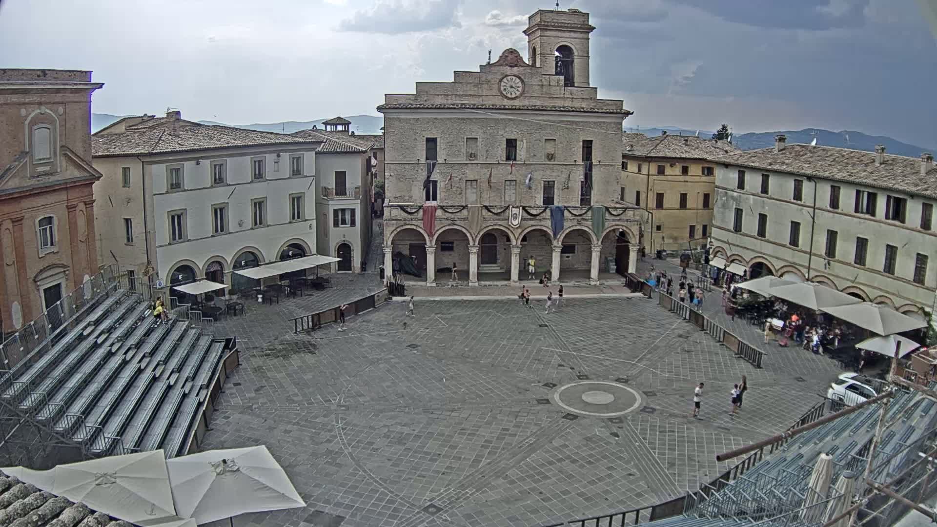 A large town square with a stone civic building at its center is surrounded by other buildings under a clear, sunny sky.