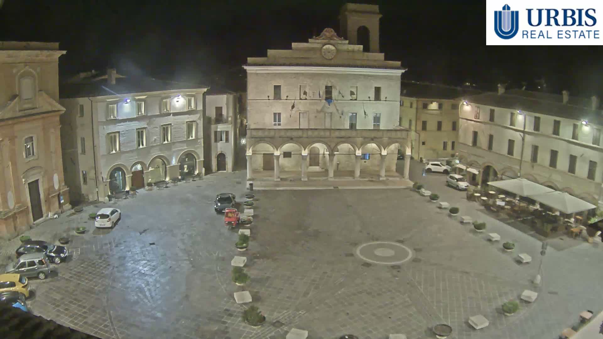A historic European town square is depicted at night under a clear sky, illuminated by streetlights, with an ornate central building featuring arches and a clock tower, a church, several parked vehicles, and an outdoor dining area on the right.