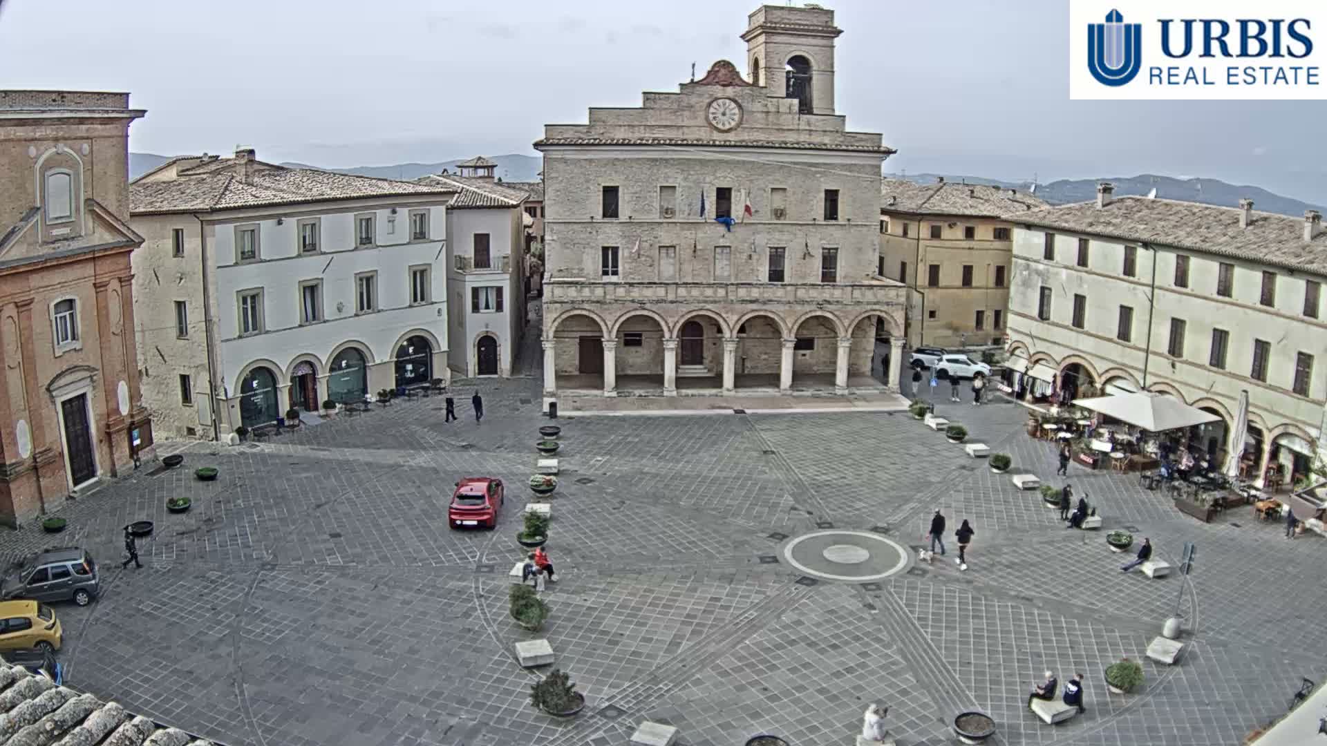 On an overcast day, a historic European town square is seen from above, featuring a prominent stone building with a clock tower and arched facade, surrounded by other multi-story buildings housing shops and outdoor cafes, with pedestrians and a few cars populating the paved plaza.