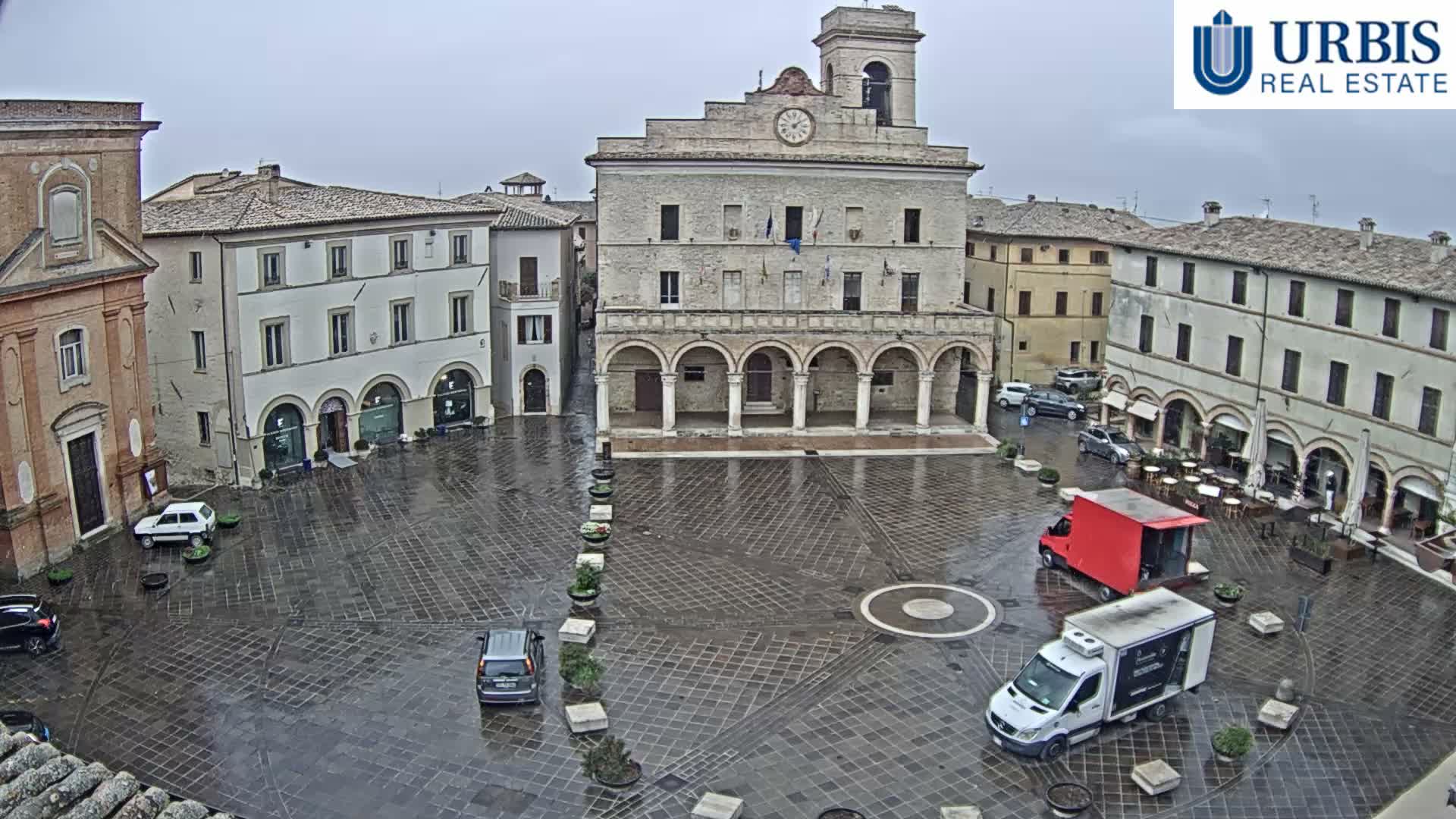 On an overcast day, a historic European town square is seen from above, featuring a prominent stone building with a clock tower and arched facade, surrounded by other multi-story buildings housing shops and outdoor cafes, with pedestrians and a few cars populating the paved plaza.