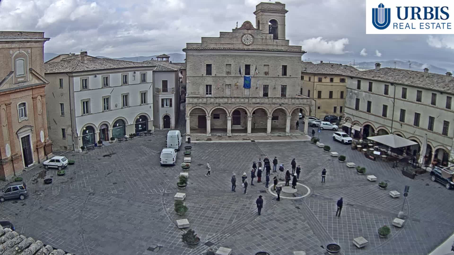 A wide shot reveals a European town square on an overcast day, surrounded by multi-story buildings, including a prominent one with a clock tower and arched entrance, with several people gathered in the center and others walking or near parked cars.
