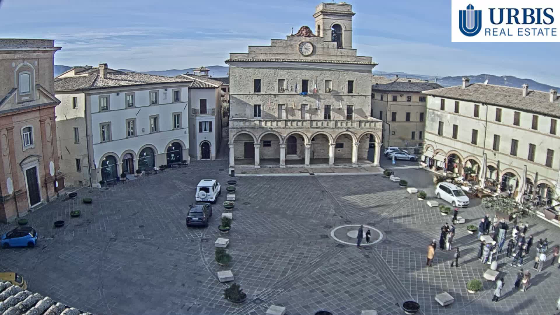 A bustling historic town square is seen on a bright, clear day, featuring a large stone building with a clock tower and arched loggia, surrounding multi-story structures with cafes, several cars, and numerous people walking and gathering on the paved ground.