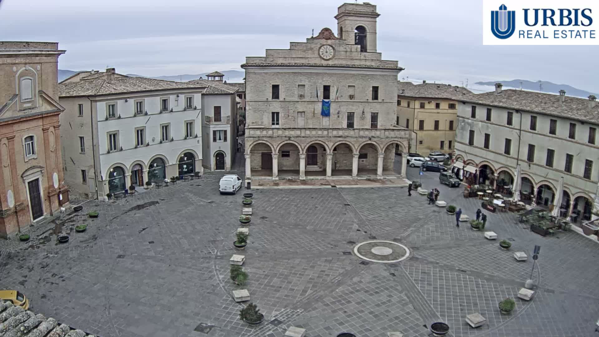 A historic European town square is bustling with scattered people, outdoor cafe seating, and parked cars, all surrounded by old stone buildings and arcades under a cloudy sky.