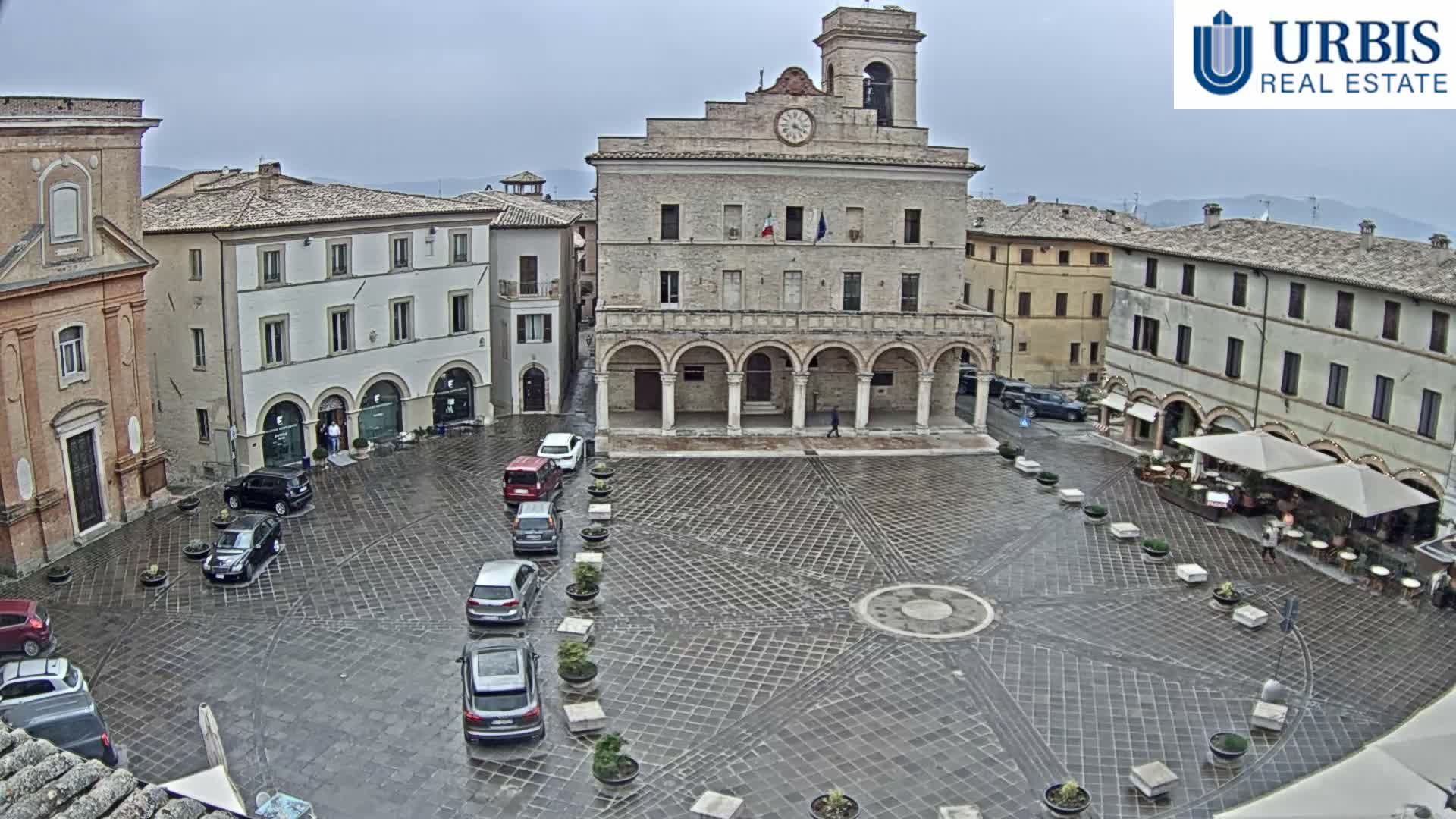 A historic European town square is bustling with scattered people, outdoor cafe seating, and parked cars, all surrounded by old stone buildings and arcades under a cloudy sky.