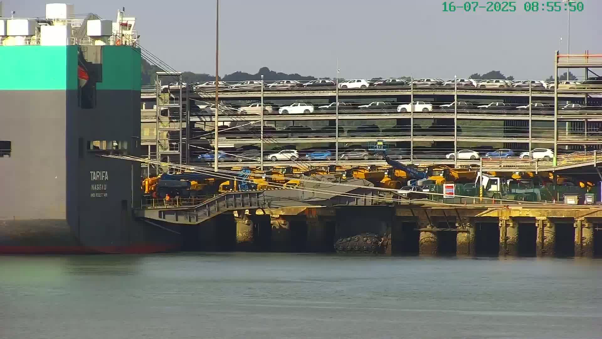 A large cargo ship is docked at a port, next to a multi-level structure filled with cars and several yellow construction vehicles.