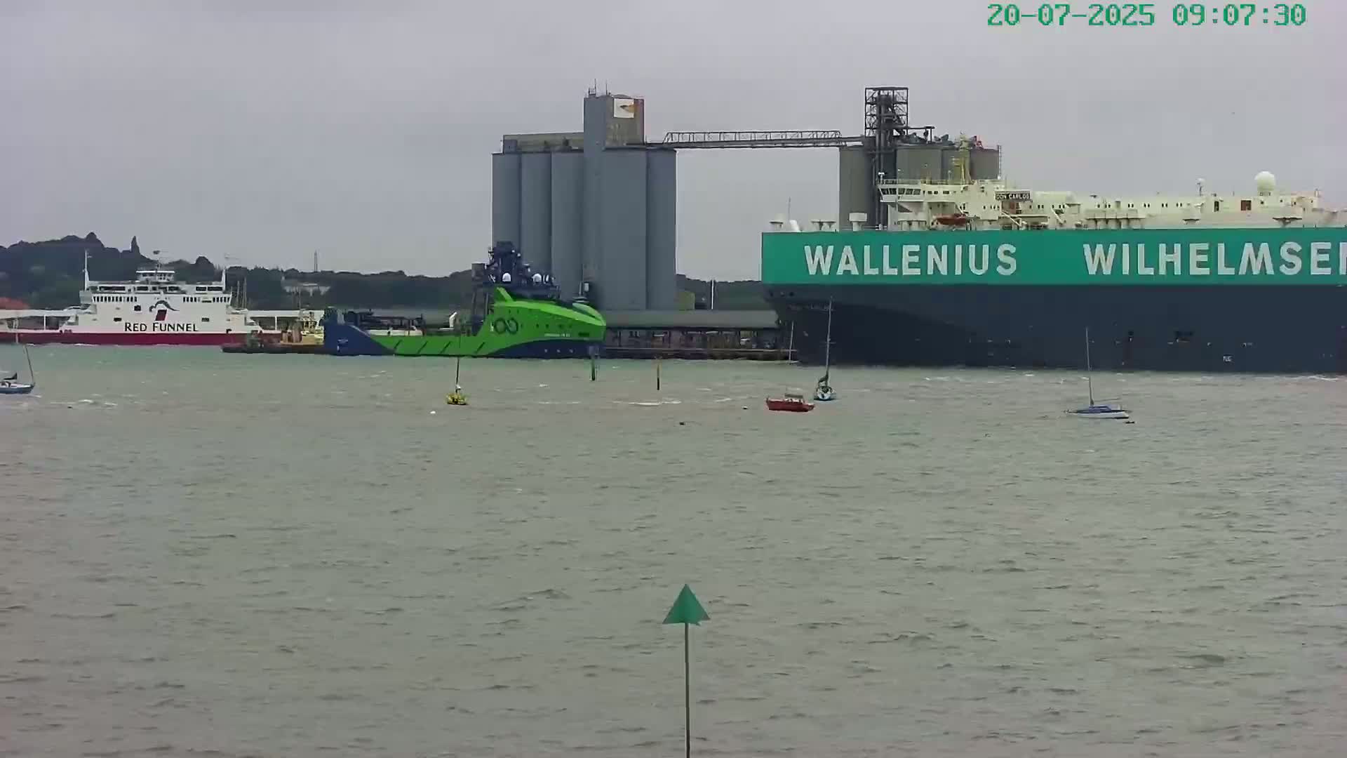 A ferry, a large green tugboat, and a large cargo ship are moored near gray grain silos in choppy gray water under an overcast sky.
