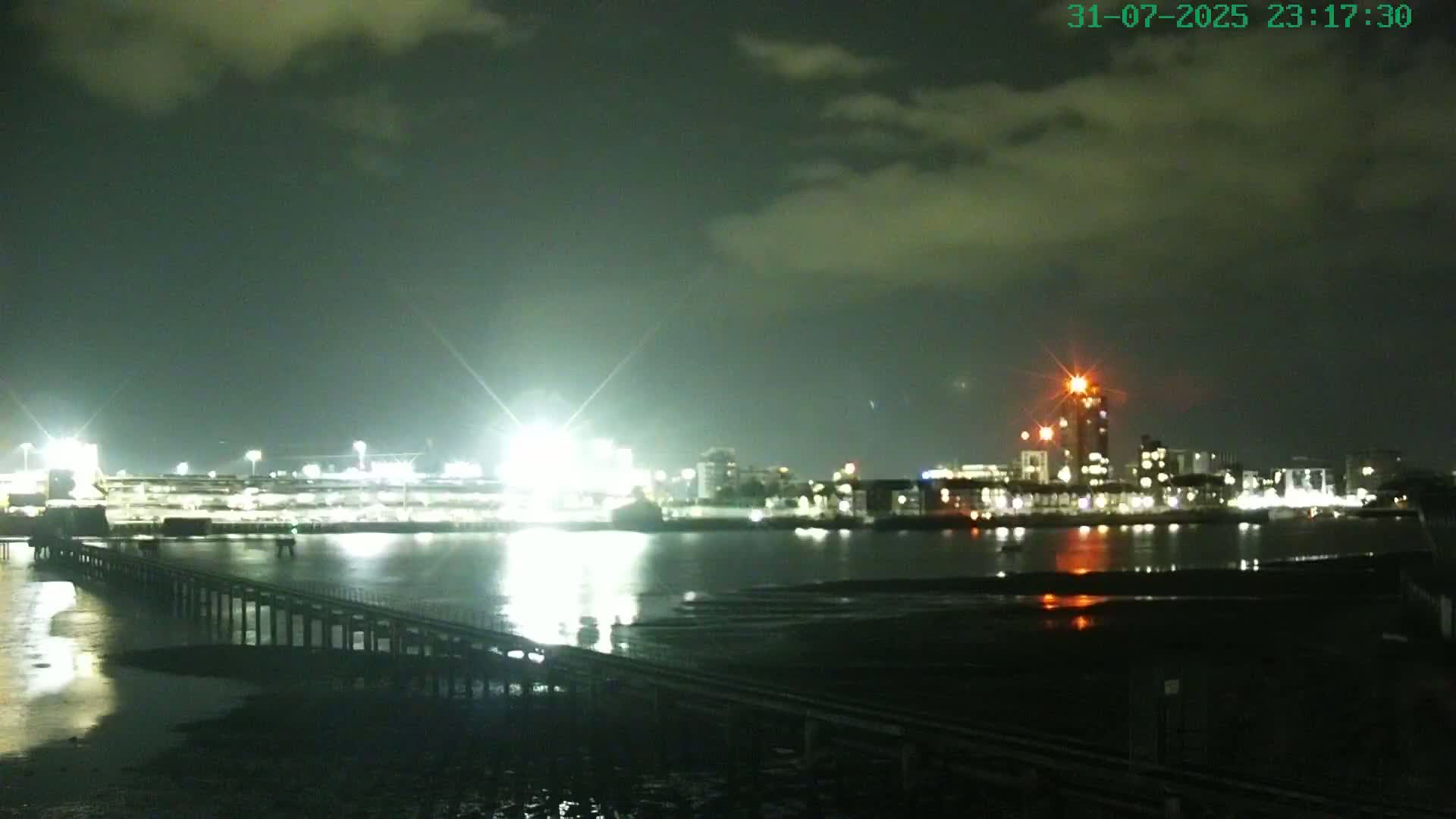 A pier extends into a calm body of water at night, with brightly lit buildings and a stadium across the water under a cloudy sky.