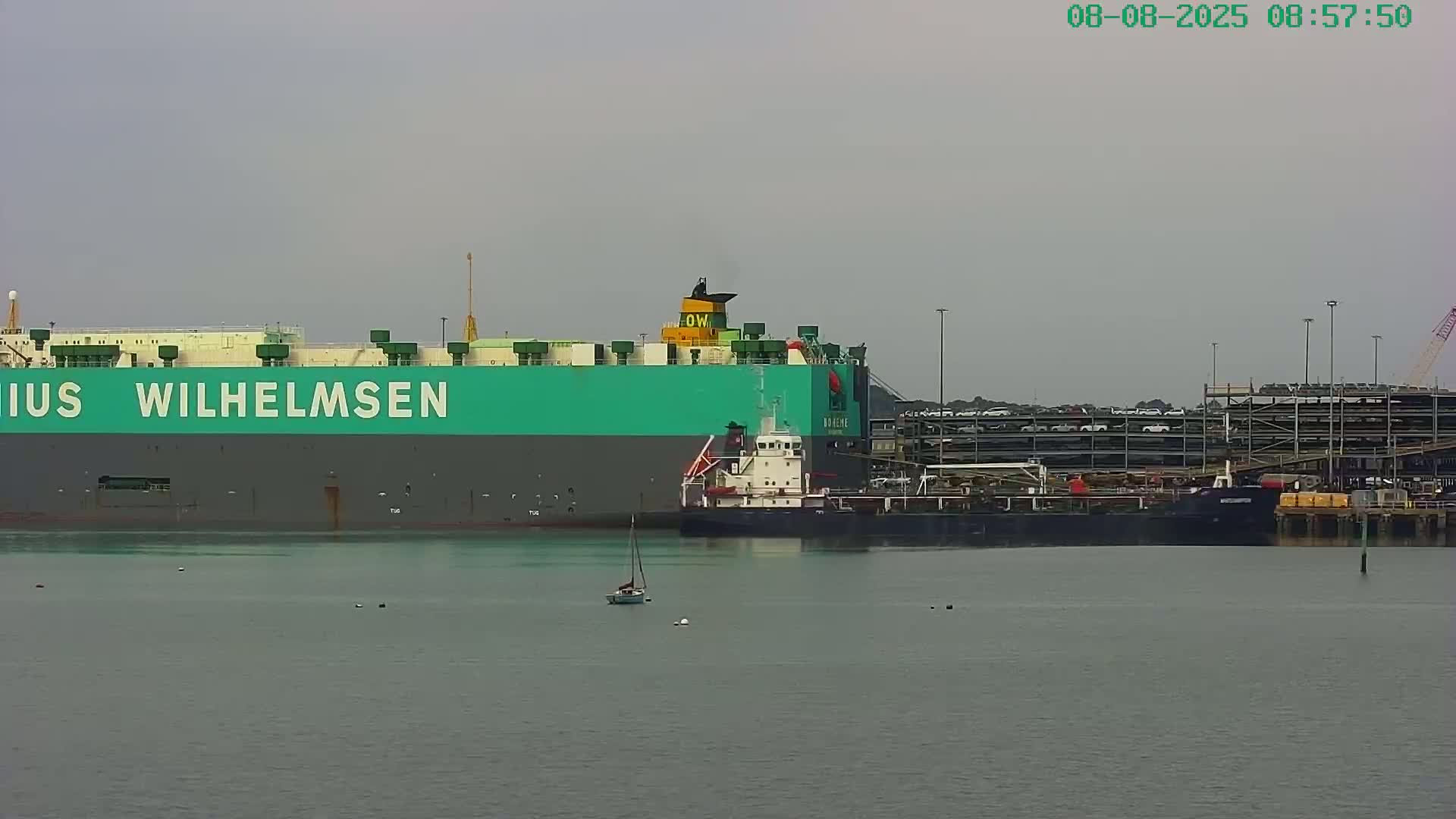 A large green and gray cargo ship is docked next to a smaller dark-colored ship in calm, overcast water, with a sailboat visible in the foreground and a multi-story car storage facility in the background.