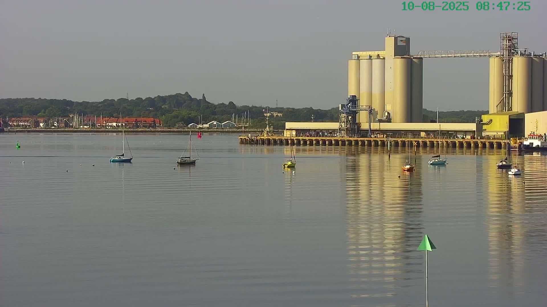 Several sailboats are moored in calm water near a large industrial facility with tall silos under a mostly clear sky.