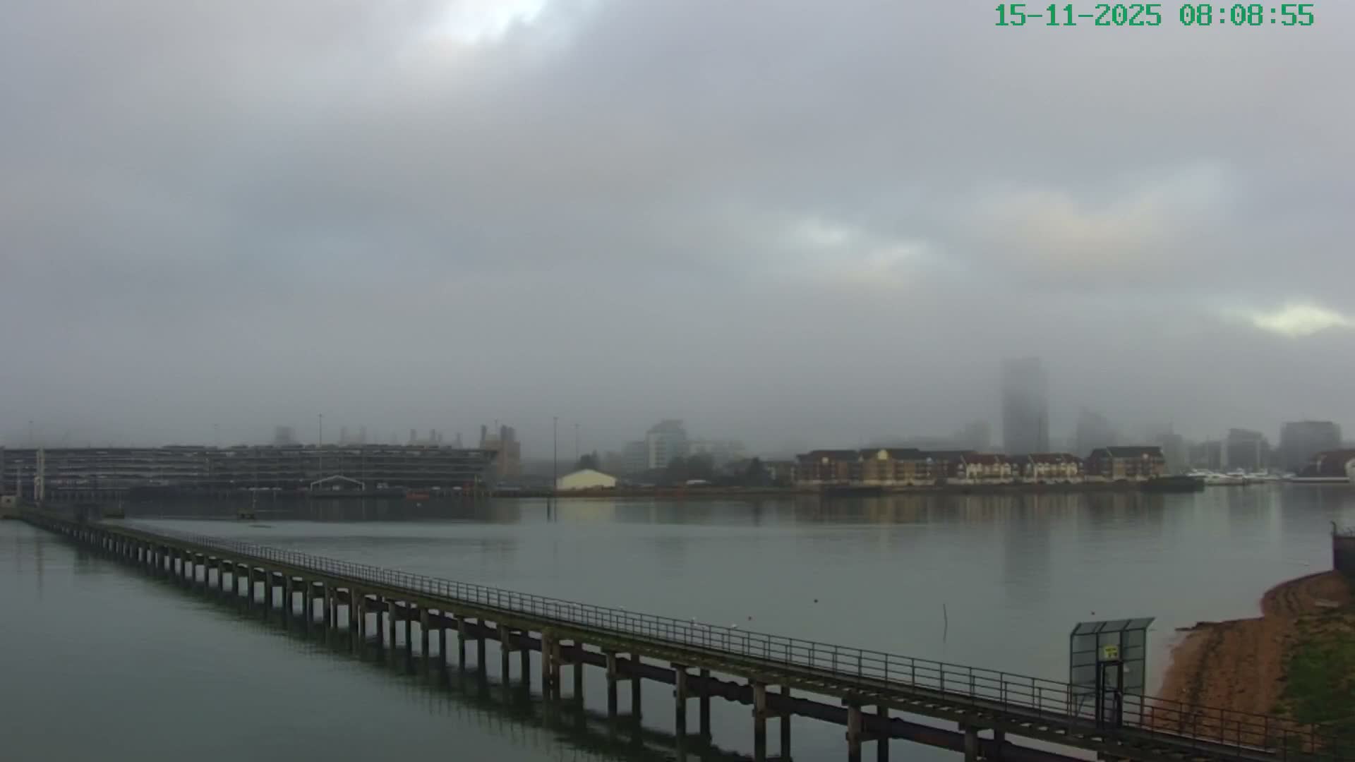 A long wooden pier extends over a calm body of water towards a cityscape with buildings shrouded in a dense, foggy atmosphere under an overcast sky.
